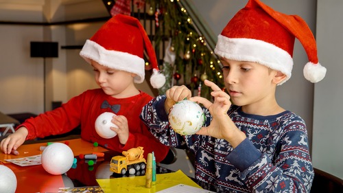 Due bambini con cappelli di Natale decorano palline natalizie seduti a un tavolo durante un’attività creativa per il calendario dell'avvento