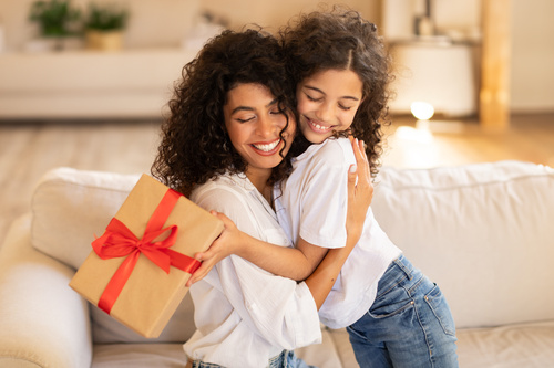 Mamma e figlia sorridenti si abbracciano sul divano mentre tengono in mano un regalo confezionato.