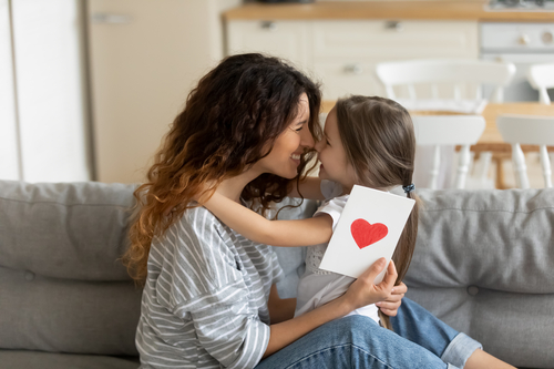 Bambina abbraccia la mamma sul divano e le regala un biglietto con un cuore rosso